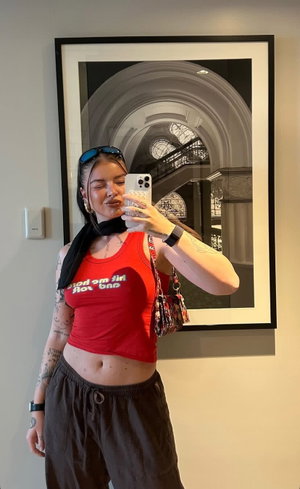 Woman in red crop top and dark pants, tattoos, mirror selfie indoors.