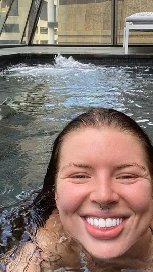 Woman with wet hair smiling in a pool, water around shoulders, outdoor setting.