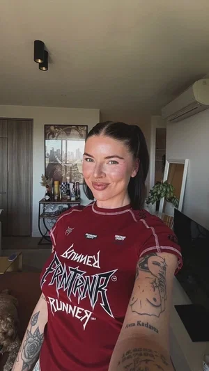 Woman with tattoos in red graphic tee, taking selfie indoors in living room.