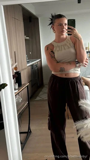 Woman with tattoos in a white crop top and dark pants, standing in a modern kitchen.