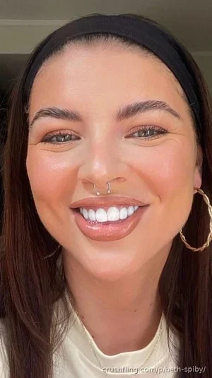 Woman with long dark hair, nose ring, hoop earrings, smiling in a close-up selfie indoors.