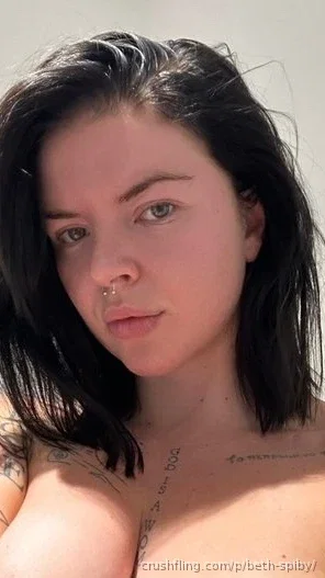 Woman with dark hair and tattoos on chest, topless, outdoors in sunlight.
