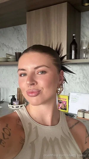 Woman with tattoos and nose ring in a beige tank top, indoors in a kitchen setting.
