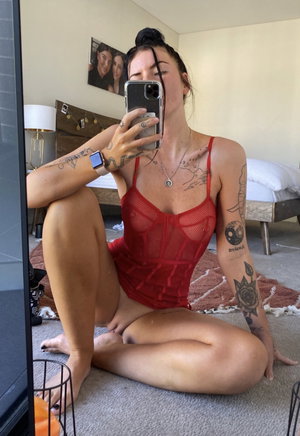 Tattooed woman in red lingerie taking mirror selfie in bedroom.