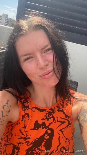 Woman with dark hair and tattoos wearing orange tank top, outdoor selfie.