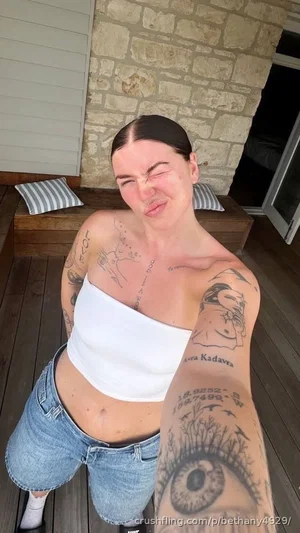 Woman with tattoos in white top and denim shorts, posing on wooden deck outdoors.