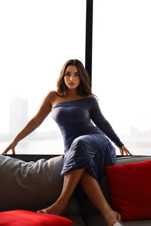 Woman in blue one-shoulder dress sitting on couch by window, indoor setting.