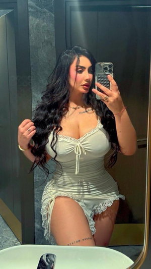 Woman with long dark hair in white lace lingerie taking mirror selfie in bathroom.