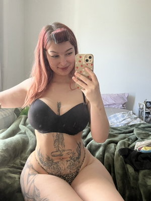 Woman with pink hair and tattoos wears black bra and leopard thong, sitting on bed in bedroom.