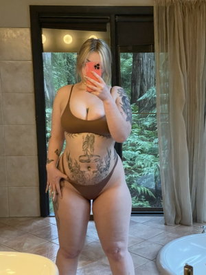Woman in brown lingerie with tattoos takes mirror selfie in bathroom.