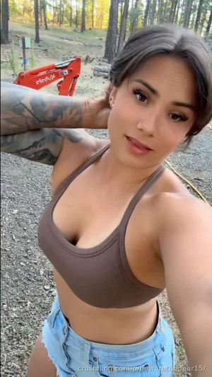 Woman in brown sports bra and denim shorts, outdoors in wooded area, tattooed arm visible.