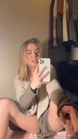 Woman with blonde hair in a gray sweater and black underwear, sitting in a bedroom taking a mirror