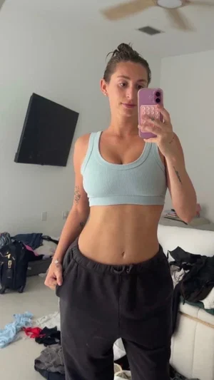 Woman in light blue crop top and black pants takes mirror selfie in messy bedroom.