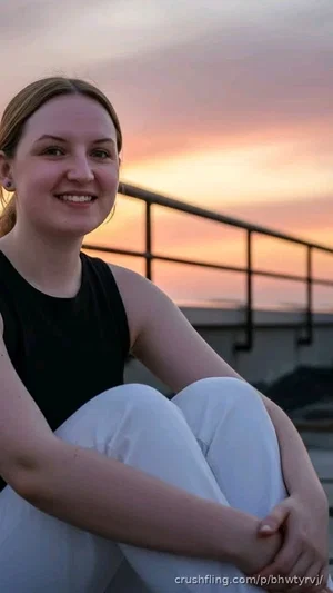 Woman in black tank top and white pants sitting outdoors at sunset.