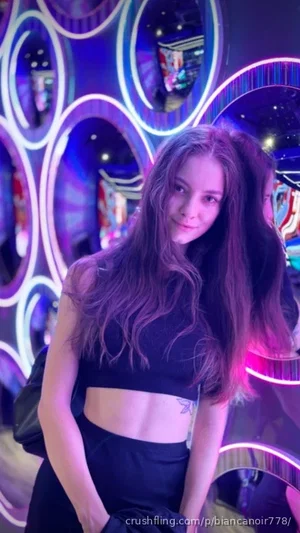 Woman with long hair in black crop top and pants, posing in neon-lit indoor setting.