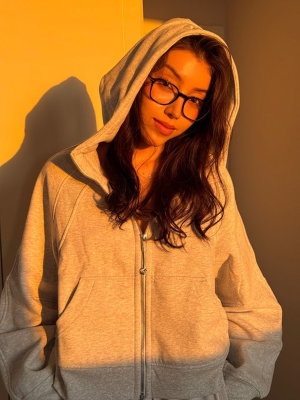Woman with long dark hair and glasses wearing a gray hoodie indoors.