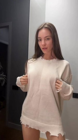 Woman in oversized cream sweater, standing indoors, pulling sleeves, long brown hair.
