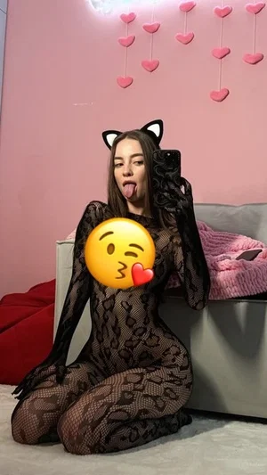 Woman in black mesh bodysuit with cat ears, kneeling on floor in pink room.