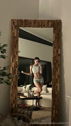 Woman in light-colored sports bra and shorts poses in mirror selfie in living room.