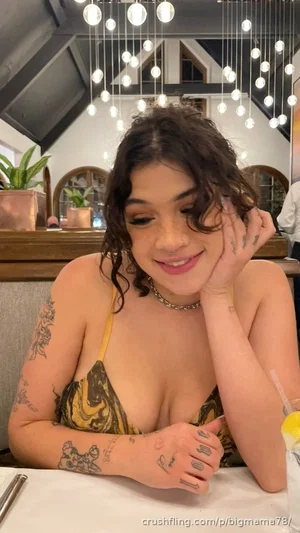 Woman with curly hair and tattoos wears a yellow patterned top, sitting at a table in a restaurant