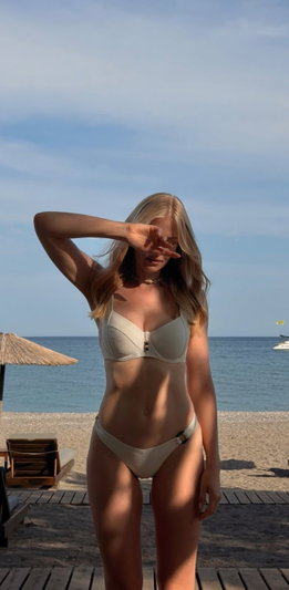 Blonde woman in beige bikini standing on beach, hand shielding eyes, ocean background.