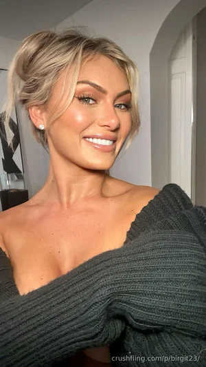 Woman with blonde hair in a bun, wearing a dark off-shoulder sweater, smiling indoors.