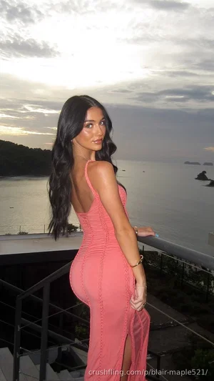 Woman in pink form-fitting dress standing on balcony overlooking water at dusk.