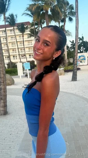 Woman in blue top and light blue skirt, smiling, outdoors near palm trees and building.