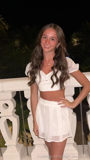 Woman with long brown hair in white crop top and skirt, standing on balcony at night.