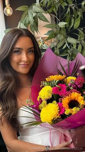 Woman with long brown hair in white top holding colorful bouquet indoors near plants.