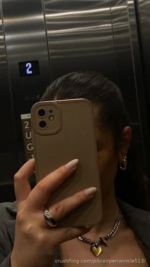 Woman with dark hair in elevator, holding phone, wearing chain necklace and ring.