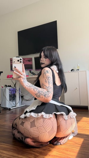 Woman with tattoos in black dress and fishnet stockings kneeling indoors, holding phone for selfie.