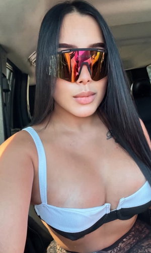 Buxom woman with long black hair in white top and lace bottoms in car.