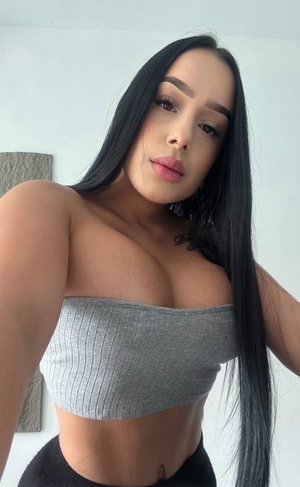 Buxom woman with long black hair in gray tube top and black leggings indoors.