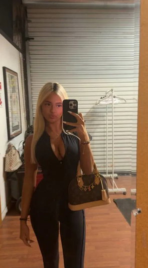 Blonde woman in black outfit taking mirror selfie indoors.