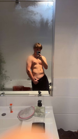 Shirtless man with towel around waist taking mirror selfie in bathroom.