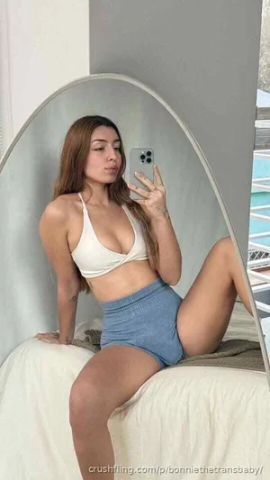 Woman in white top and blue shorts posing on bed in front of mirror.