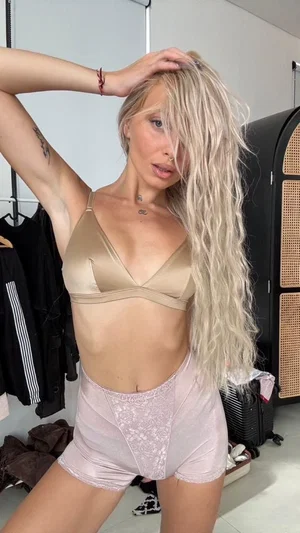 Blonde woman in beige bra and pink lace shorts posing indoors.