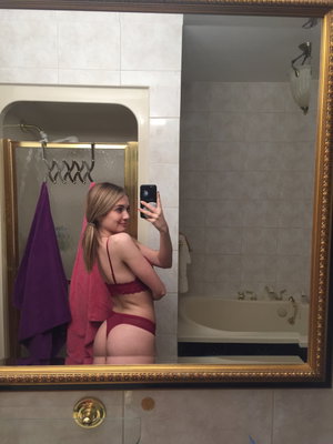 Woman in red lingerie takes mirror selfie in bathroom with tiled walls and bathtub.