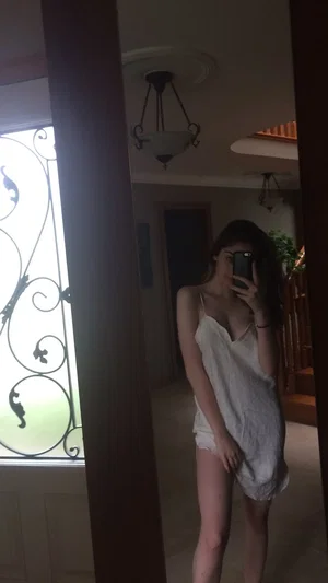 Woman in white slip dress taking mirror selfie in dimly lit indoor space.