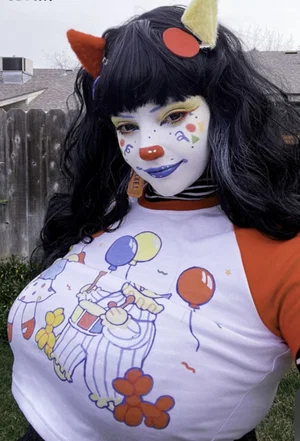 Woman with clown makeup and black wig wearing a colorful graphic tee outdoors.