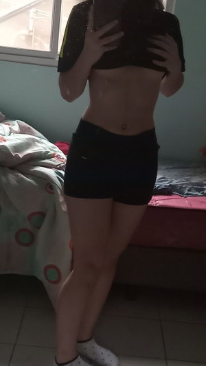 Slim woman topless in shorts, bedroom mirror selfie.