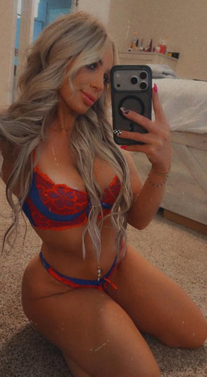 Blonde woman in red and blue lingerie taking bedroom selfie.