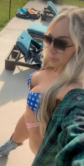 Blonde woman in star-patterned bikini top and plaid shirt, standing outdoors near lounge chairs.