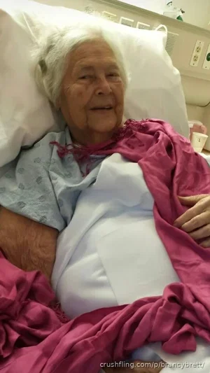 Elderly woman in hospital bed, covered with pink blanket, wearing hospital gown.