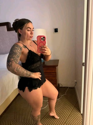 Woman with tattoos in black lace lingerie posing in bedroom mirror selfie.