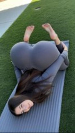 Woman in gray leggings lying on yoga mat on grass, long dark hair, outdoor setting.