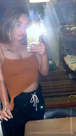Woman with blonde hair in orange crop top and black pants, taking mirror selfie in indoor setting.