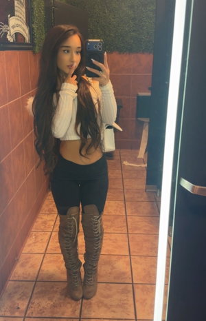 Asian woman in white crop top, black leggings, brown boots in bathroom.