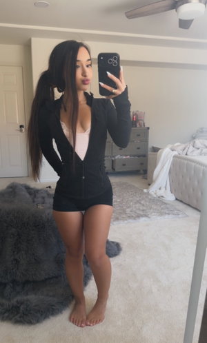 Asian woman in open black jacket, lace bra, shorts in bedroom.